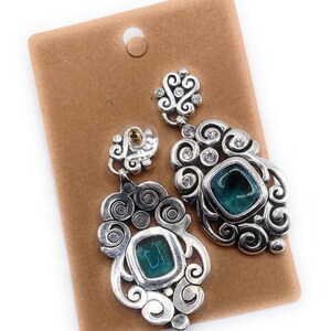 May include: Silver-toned statement earrings with a teal square stone and clear crystals. The earrings have a decorative, swirling design and are displayed on a brown earring card.
