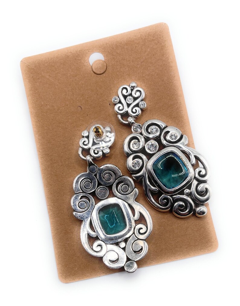 May include: Silver-toned statement earrings with a teal square stone and clear crystals. The earrings have a decorative, swirling design and are displayed on a brown earring card.
