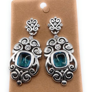 May include: A pair of silver-toned drop earrings with a decorative design. Each earring features a square, teal-colored stone surrounded by ornate swirls and clear crystals. The earrings are displayed on a brown card.