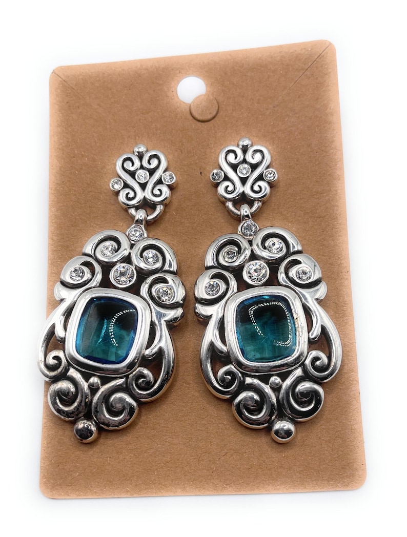 May include: A pair of silver-toned drop earrings with a decorative design. Each earring features a square, teal-colored stone surrounded by ornate swirls and clear crystals. The earrings are displayed on a brown card.