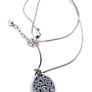 K&ouml;nnte beinhalten: Silberfarbene Halskette mit einem Anh&auml;nger mit einem wirbelnden, durchbrochenen Design. Der Anh&auml;nger ist tropfenf&ouml;rmig und hat einen kleinen gr&uuml;nen Akzent. Die Halskette hat eine zarte Kette und einen verstellbaren Verschluss.