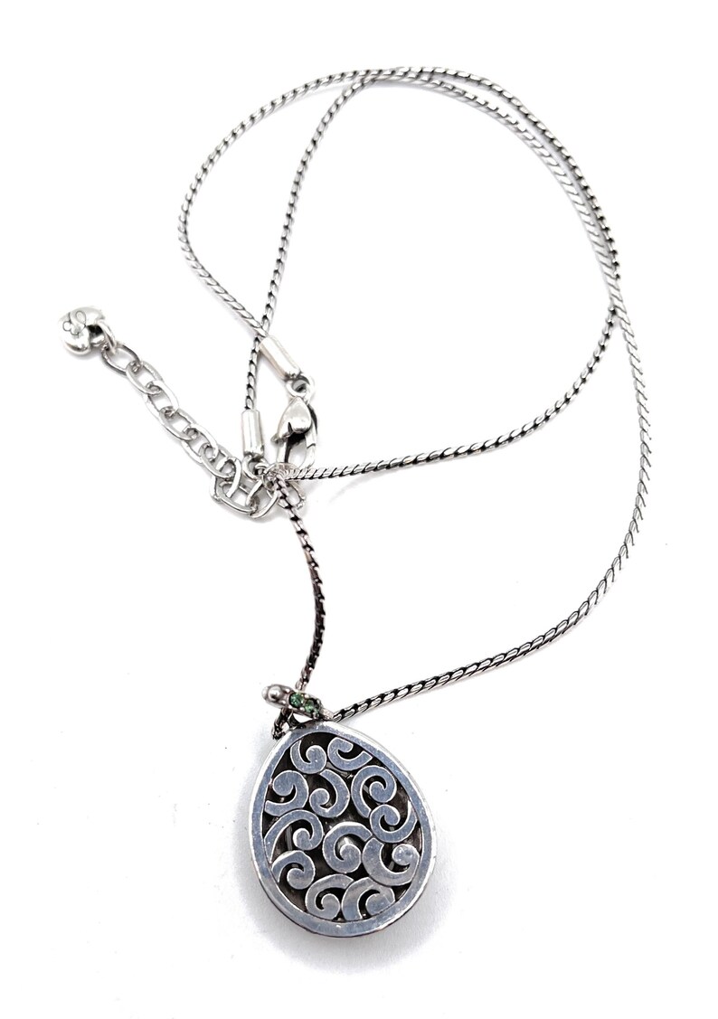 K&ouml;nnte beinhalten: Silberfarbene Halskette mit einem Anh&auml;nger mit einem wirbelnden, durchbrochenen Design. Der Anh&auml;nger ist tropfenf&ouml;rmig und hat einen kleinen gr&uuml;nen Akzent. Die Halskette hat eine zarte Kette und einen verstellbaren Verschluss.
