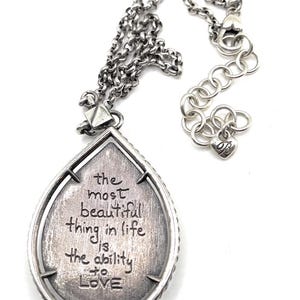 May include: Silver-toned necklace with a teardrop pendant. The pendant features the engraved quote, "the most beautiful thing in life is the ability to LOVE." The necklace has a chain with an adjustable length.