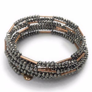 May include: A wrap bracelet featuring multiple strands of faceted, gray beads. The beads are strung together with copper-colored metal bars. A small, gold-colored charm is attached. The bracelet is designed to wrap around the wrist.