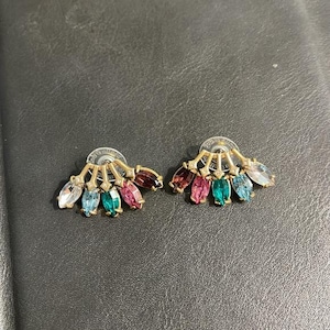 Pode incluir: Um par de brincos de pressão dourados. Cada brinco apresenta um design em forma de leque com pedras preciosas coloridas em forma de marquise em tons de transparente, azul, verde, rosa e castanho. Os brincos estão sobre um fundo escuro.