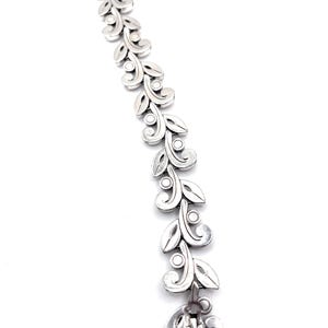 Puede incluir: Una pulsera plateada con un dise&ntilde;o repetido de hojas y espirales. La pulsera presenta una serie de hojas estilizadas conectadas por peque&ntilde;os c&iacute;rculos. El cierre es un elemento decorativo con un motivo de hoja a juego. La pulsera se muestra sobre un fondo blanco.