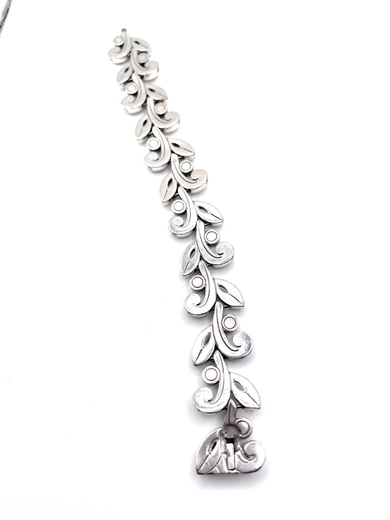 Puede incluir: Una pulsera plateada con un dise&ntilde;o repetido de hojas y espirales. La pulsera presenta una serie de hojas estilizadas conectadas por peque&ntilde;os c&iacute;rculos. El cierre es un elemento decorativo con un motivo de hoja a juego. La pulsera se muestra sobre un fondo blanco.