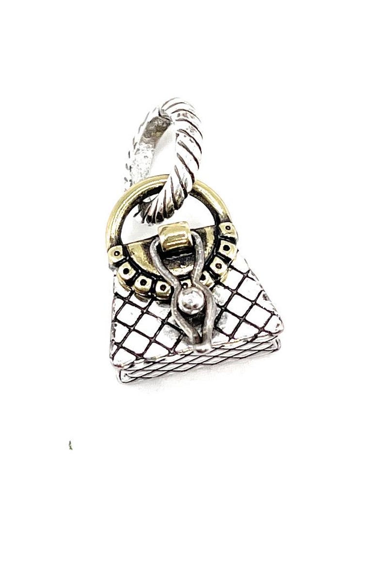 May include: A silver-toned charm in the shape of a handbag, featuring a textured, diamond-patterned body. The charm has a gold-colored handle and clasp, and a twisted silver loop for attachment. A small, silver ball accents the front.