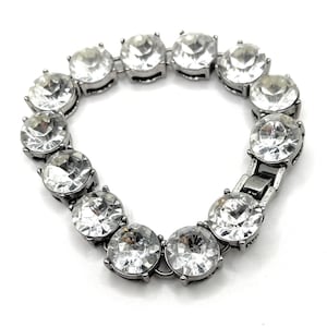 Può includere: Un bracciale argentato con grandi gemme rotonde e trasparenti. Le gemme sono incastonate in una montatura metallica e collegate per formare una catena flessibile. Il bracciale ha una chiusura a fermaglio.