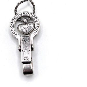 Puede incluir: Colgante plateado con un dise&ntilde;o en forma de coraz&oacute;n dentro de un marco circular adornado con recortes en forma de coraz&oacute;n. El colgante tiene un cierre de bisagra y una cadena trenzada para colgar. El coraz&oacute;n tiene remolinos decorativos.
