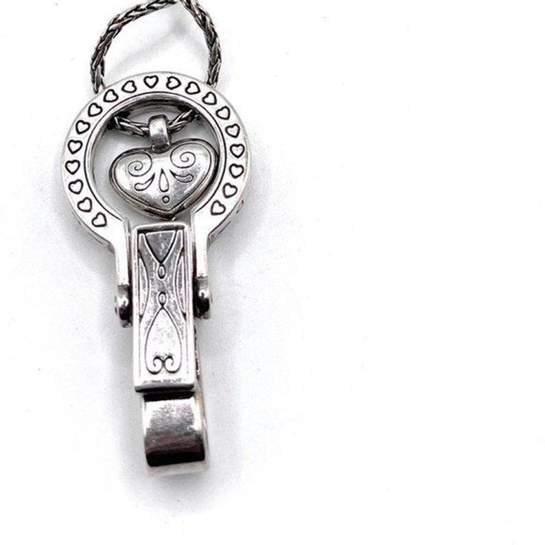 Puede incluir: Colgante plateado con un dise&ntilde;o en forma de coraz&oacute;n dentro de un marco circular adornado con recortes en forma de coraz&oacute;n. El colgante tiene un cierre de bisagra y una cadena trenzada para colgar. El coraz&oacute;n tiene remolinos decorativos.