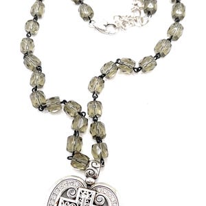 Peut inclure: Un collier avec un pendentif en forme de c&oelig;ur orn&eacute; d'une croix et de cristaux transparents. La cha&icirc;ne est compos&eacute;e de perles facett&eacute;es gris fum&eacute;. Le fermoir est argent&eacute;.