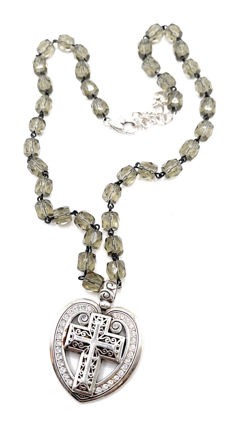 Peut inclure: Un collier avec un pendentif en forme de c&oelig;ur orn&eacute; d'une croix et de cristaux transparents. La cha&icirc;ne est compos&eacute;e de perles facett&eacute;es gris fum&eacute;. Le fermoir est argent&eacute;.