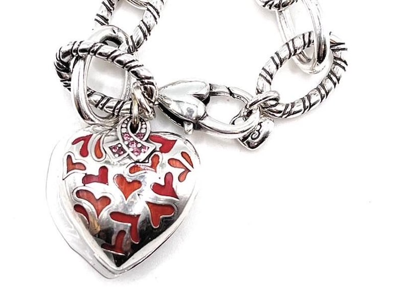 May include: Silver charm bracelet with a heart-shaped charm featuring a red and white enamel design. The charm also has a pink ribbon with small pink rhinestones.