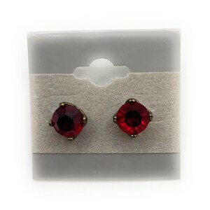 May include: A pair of stud earrings featuring square, faceted, deep red gemstones set in antique gold-tone metal. The earrings are displayed on a gray earring card with a neutral background.