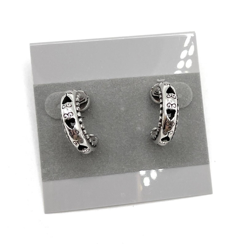 May include: A pair of silver-toned hoop earrings. Each earring has a curved design with heart-shaped cutouts and decorative detailing. The earrings are displayed on a gray earring card.