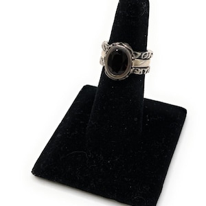 May include: A silver ring with an oval, dark gemstone. The ring has a decorative band with etched patterns. The ring is displayed on a black velvet ring holder against a white background.