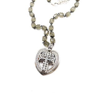 Peut inclure: Un collier argent&eacute; avec un pendentif en forme de c&oelig;ur orn&eacute; d'une croix. La croix pr&eacute;sente des motifs complexes et est entour&eacute;e de petites pierres transparentes. Le collier est compos&eacute; de perles facett&eacute;es gris fum&eacute;.