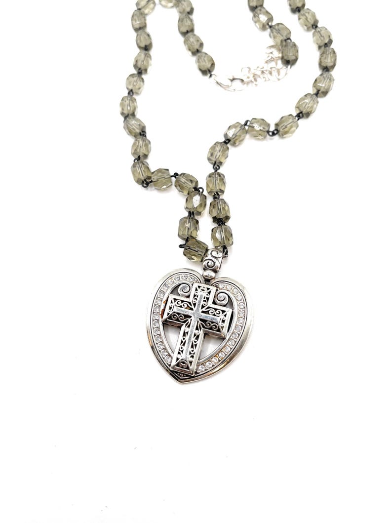 Peut inclure: Un collier argent&eacute; avec un pendentif en forme de c&oelig;ur orn&eacute; d'une croix. La croix pr&eacute;sente des motifs complexes et est entour&eacute;e de petites pierres transparentes. Le collier est compos&eacute; de perles facett&eacute;es gris fum&eacute;.