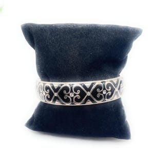 May include: A silver-toned metal bangle bracelet featuring a repeating black enamel scrollwork pattern. The design is accented with small, clear, round inset stones placed at intervals along the center of the black enamel sections.