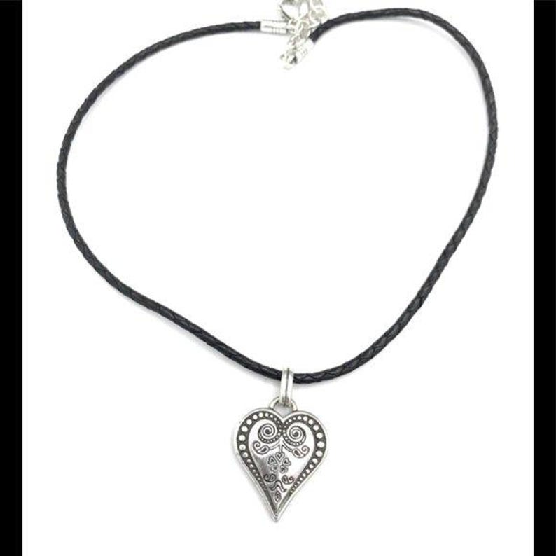 K&ouml;nnte beinhalten: Halskette mit silbernem Herzanh&auml;nger und schwarzem Band. Der Herzanh&auml;nger hat ein verziertes Design mit Wirbeln und Punkten. Die Halskette befindet sich auf wei&szlig;em Hintergrund.