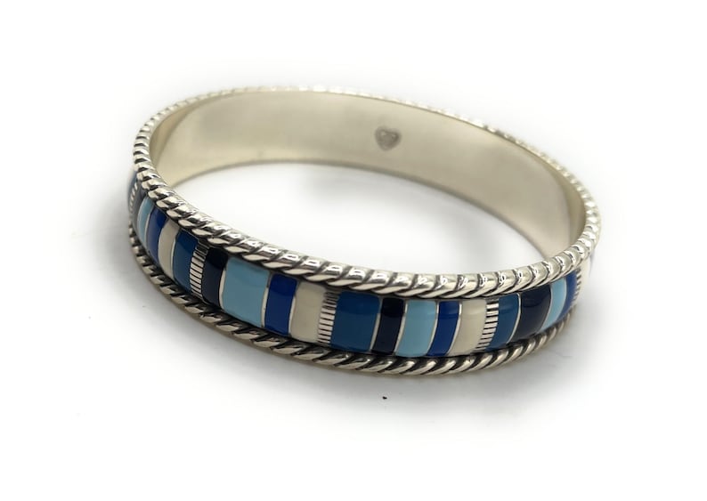 Może przedstawiać: Srebrna bransoletka typu obręcz z paskami w odcieniach niebieskiego i białego. Bransoletka ma plecioną krawędź i gładkie, wypolerowane wnętrze. Wewnątrz bransoletki widoczny jest znak w kształcie serca.