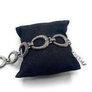 May include: Silver-toned bracelet featuring a series of ornate, open oval links with a detailed scroll design. The bracelet is displayed on a black pillow against a white background, showcasing its intricate craftsmanship.