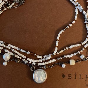 May include: A multi-strand necklace with white and brown beads, a silver chain, and a large, round, iridescent pendant. The necklace is on a brown surface with the text "Silpad find your" visible.