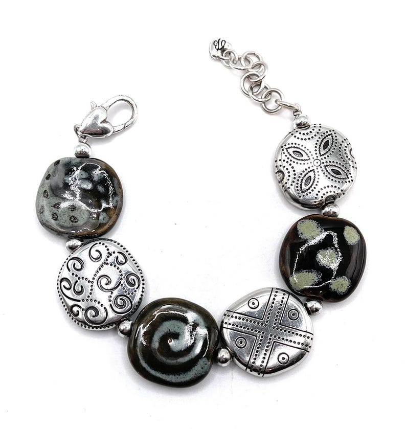 Puede incluir: Un brazalete de plata con cinco dijes. Dos de los dijes son negros y blancos con un patr&oacute;n de espiral. Los otros tres dijes son de plata con un dise&ntilde;o decorativo.