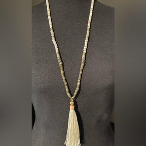 May include: A long beaded necklace featuring a mix of light-colored, translucent, and dark olive-green square beads separated by small metallic spacers. The necklace terminates in a long, light beige fabric tassel attached with a metallic cap.