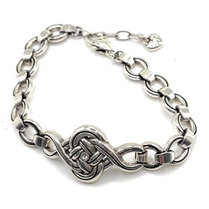 May include: Silver-toned bracelet featuring a central knot design with a small, clear gemstone. The bracelet has a chain-link style with adjustable length and a heart-shaped charm at the end. A piece of jewelry.