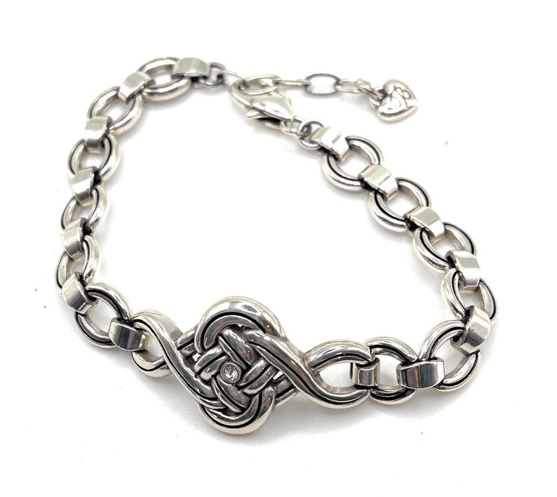 May include: Silver-toned bracelet featuring a central knot design with a small, clear gemstone. The bracelet has a chain-link style with adjustable length and a heart-shaped charm at the end. A piece of jewelry.