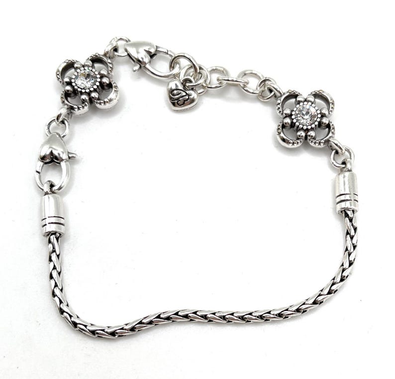 May include: A silver chain bracelet with two decorative flower charms, each with a clear rhinestone in the center. The bracelet has a heart-shaped clasp.