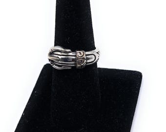 May include: A silver-colored ring with a buckle design, displayed on a black ring holder. The ring has a detailed, textured appearance with a small, decorative element in the center.