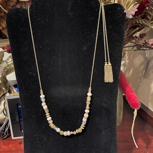 May include: A long gold-tone chain necklace featuring a variety of small beads in shades of cream, pale pink, and translucent clear, accented with metallic spacers. The necklace ends in two delicate gold-tone chain tassels hanging from the chain.