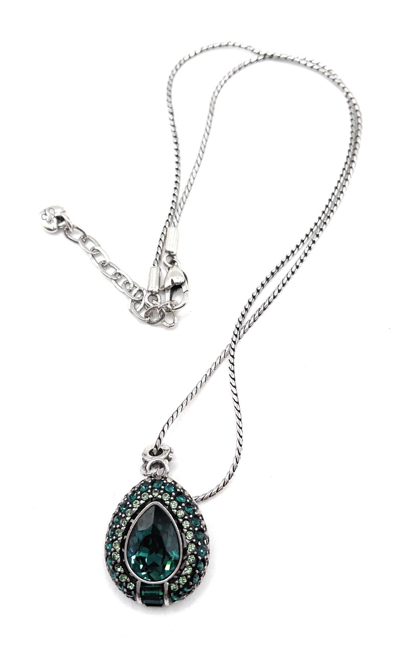 K&ouml;nnte beinhalten: Silberfarbene Halskette mit einem Anh&auml;nger, der einen gro&szlig;en, tropfenf&ouml;rmigen, dunkelgr&uuml;nen Edelstein in der Mitte hat. Der Anh&auml;nger ist von kleineren, hellgr&uuml;nen Edelsteinen umgeben. Die Kette hat ein filigranes Design und einen Karabinerverschluss.