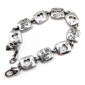 May include: Silver bracelet with square links, each with a heart-shaped cutout. The links are engraved with words such as "Love" and "Amour." A small heart charm hangs near the clasp. The bracelet has a lobster claw clasp.