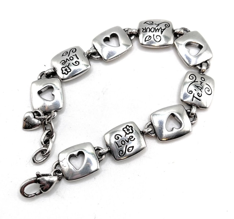May include: Silver bracelet with square links, each with a heart-shaped cutout. The links are engraved with words such as "Love" and "Amour." A small heart charm hangs near the clasp. The bracelet has a lobster claw clasp.