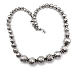 May include: A silver necklace with a series of textured, spherical beads of varying sizes. The necklace has a lobster clasp closure and a small charm near the clasp. The beads have a hammered appearance.