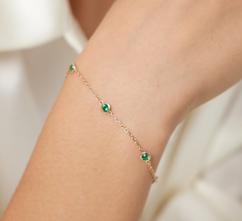 Puede incluir: Un delicado brazalete de cadena dorada con peque&ntilde;as piedras preciosas verdes. El brazalete est&aacute; en la mu&ntilde;eca de una persona.