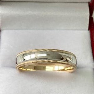 May include: A two-tone wedding band with a polished silver centre and a gold-coloured beaded edge. The ring is set in a white jewellery box with a red trim.