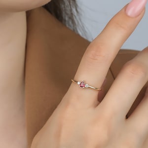 May include: A delicate gold ring with a teardrop-shaped red gemstone and two smaller white gemstones.
