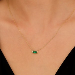 May include: A delicate gold necklace featuring a rectangular, emerald-green gemstone pendant. The pendant is set in a gold bezel and hangs from a fine gold chain. The necklace is displayed against a neutral background.
