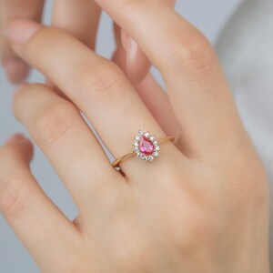May include: A gold ring featuring a pear-shaped pink gemstone set within a halo of smaller clear gemstones.