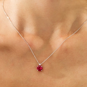 May include: A delicate silver necklace featuring a vibrant, round, red gemstone pendant. A small, clear accent stone sits above the main gem. The necklace is displayed against a neutral background, highlighting the jewelry's elegance.