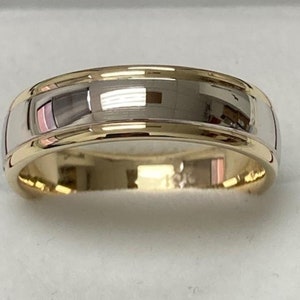 May include: A two-tone wedding band with a polished finish. The ring has a gold-coloured interior and edges, with a silver-coloured centre band. The design is simple and elegant, reflecting light.