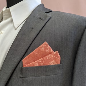 May include: A dark gray suit jacket with a pocket square. The pocket square is a folded piece of fabric with a floral pattern in shades of pink and orange.