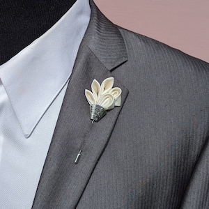 May include: A white satin flower lapel pin with a silver metal base. The pin is attached to a grey suit jacket.