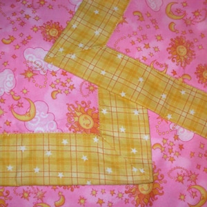 May include: A pink fabric blanket featuring a repeating pattern of yellow suns, crescent moons, and white clouds. The blanket is finished with a wide, yellow plaid border that is decorated with small white stars throughout the fabric design.