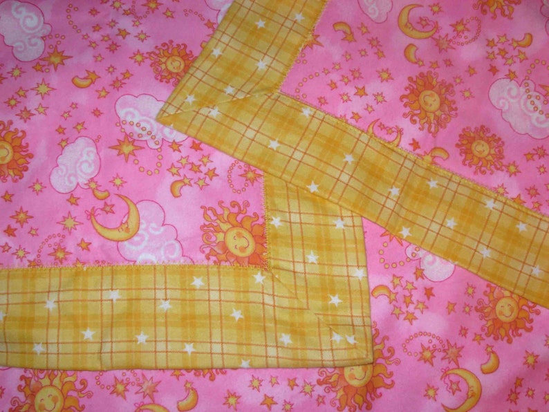 May include: A pink fabric blanket featuring a repeating pattern of yellow suns, crescent moons, and white clouds. The blanket is finished with a wide, yellow plaid border that is decorated with small white stars throughout the fabric design.
